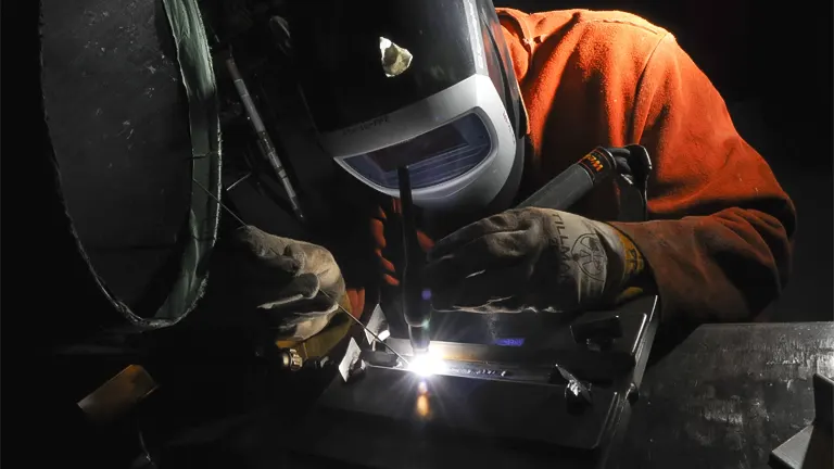 A welder in protective gear, working on a metal piece in a dark setting, illuminated by the bright light of the welding process