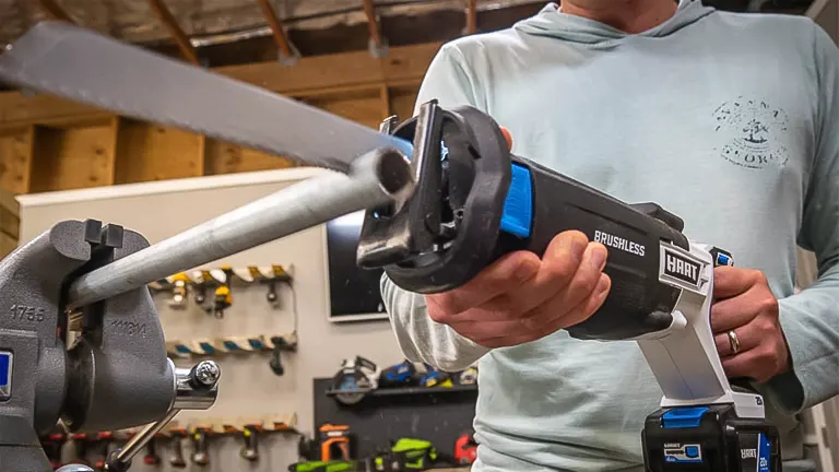 Person using a HART 20-Volt Reciprocating Saw to cut a metal pipe clamped in a vise, with a tool rack in the background