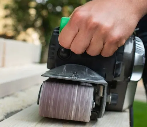 Hand holding a black and green belt sander on a wooden floor