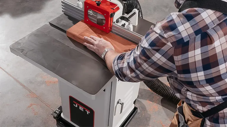 Person in plaid shirt using the white and red JET Shaper JWS-22CS Shaper on a piece of wood in a workshop