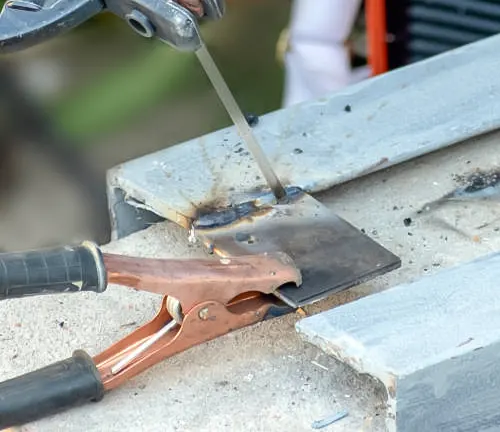 A welding clamp holding two pieces of metal together during welding, with sparks and smoke visible