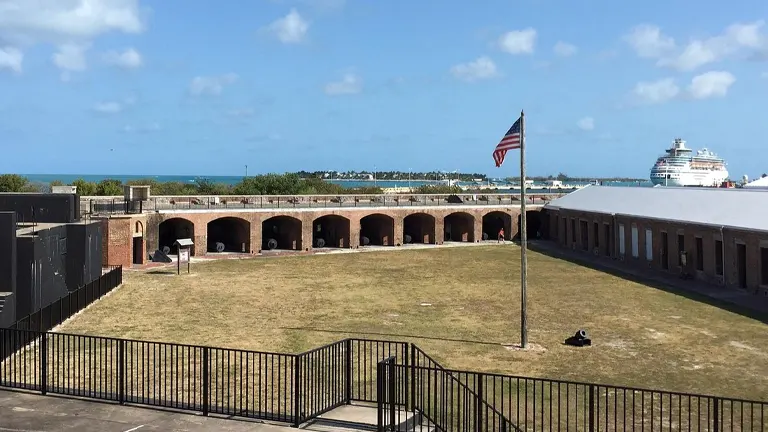 A panoramic view of Fort Zachary Taylor Historic State Park with a grassy courtyard, arched walkways, and an American flag, overlooking the ocean and a cruise ship