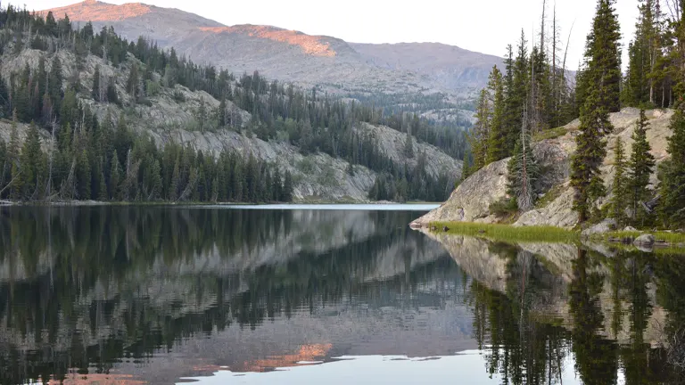 Tranquil lake reflecting the rugged, forested landscape of Cloud Peak Wilderness at dusk