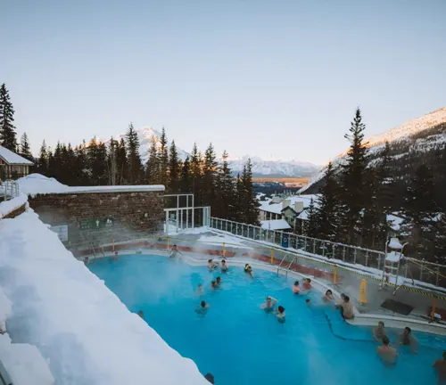 People enjoying a steaming outdoor pool in a snowy landscape with mountains
