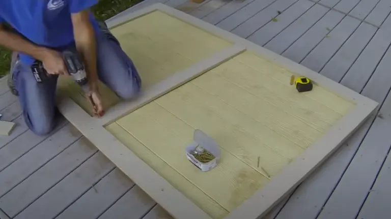 A person using a cordless drill to assemble a wooden frame on a deck, with a tape measure and screws nearby.

