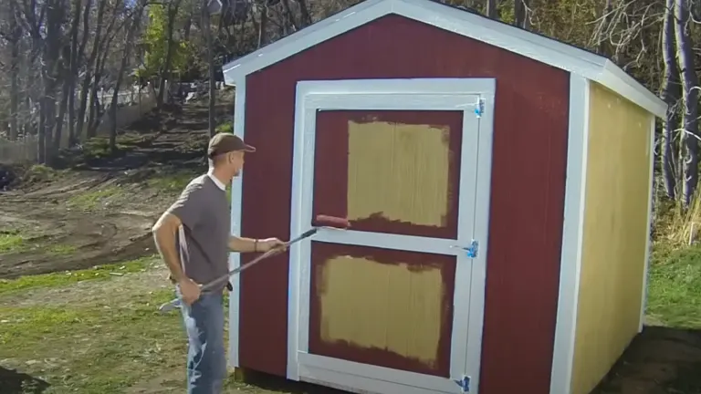 A person using a roller to paint a small, red and white shed outdoors, with trees in the background.


