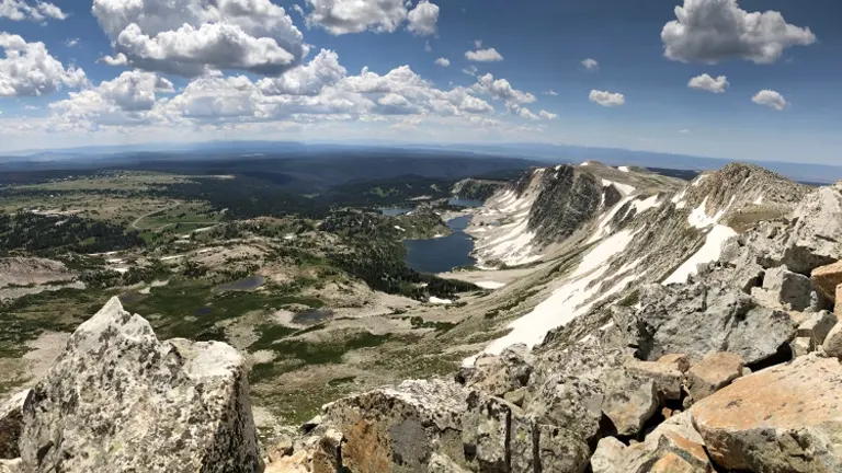 a panoramic view of Medicine Bow Peak, featuring snow-capped mountains, a clear lake, and a rocky foreground under a cloudy sky