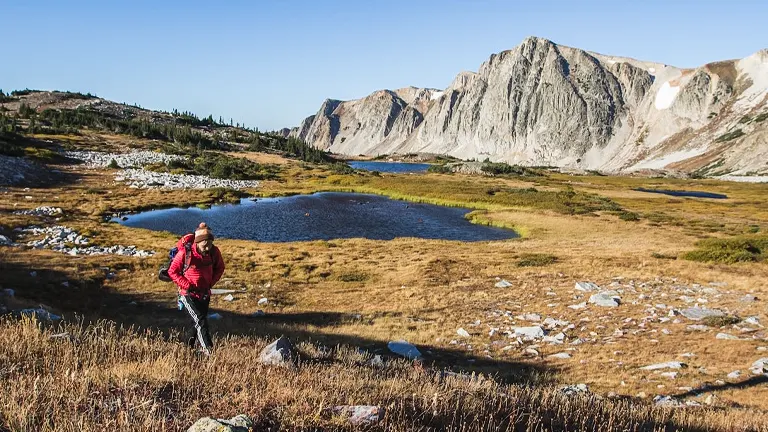 a hiker in a red jacket trekking through a vast, mountainous landscape dotted with clear blue ponds