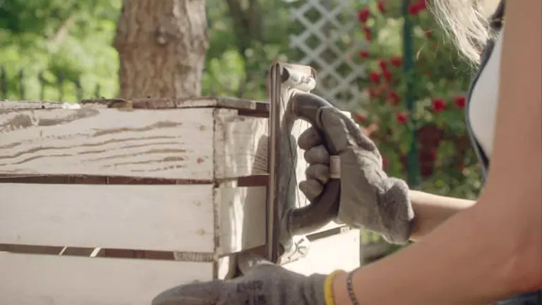 Close-up of a person using a sander on the corner of a white wooden box outdoors.