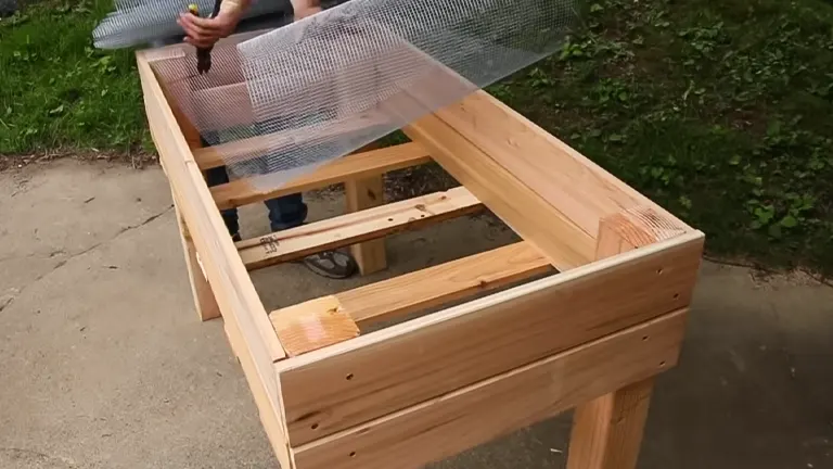 A person attaching mesh to a wooden raised garden bed frame outdoors.