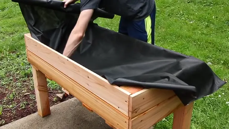 A man fitting a black liner inside a wooden raised garden bed in a grassy area.