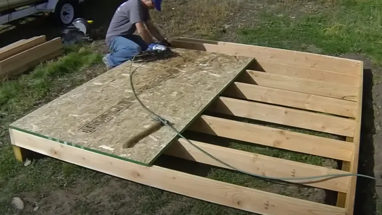 A person securing plywood on top of a nearly completed wooden raised garden bed outdoors.

