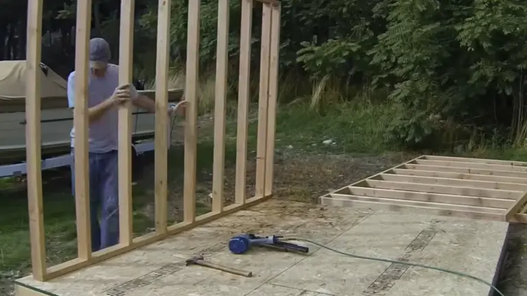 A person assembling a vertical wooden frame structure next to a completed raised garden bed, with tools scattered on plywood flooring.

