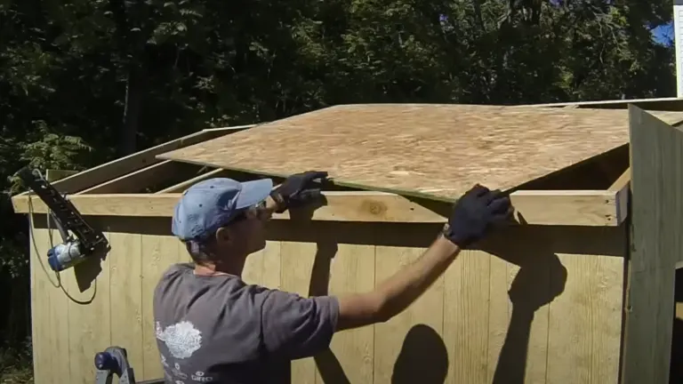 A person installing plywood on the roof of a wooden structure outdoors, with tools hanging on the wall nearby.


