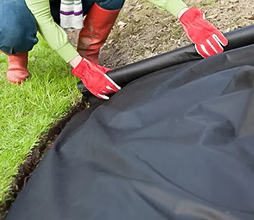 Person installing black landscape fabric on garden soil, wearing red gloves