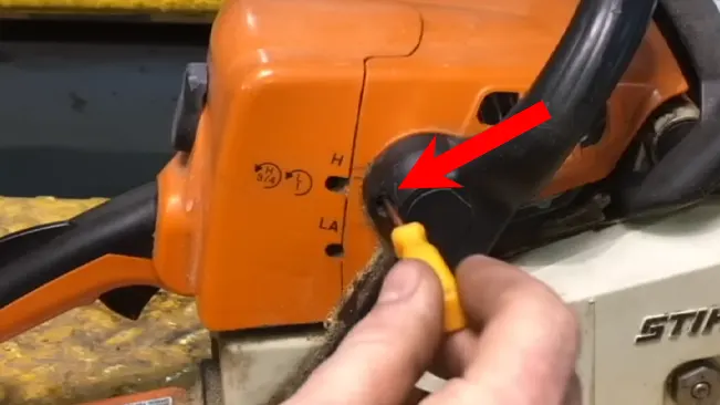 Close-up of a person's hand adjusting a STIHL chainsaw, focusing on the tensioning area indicated by the red arrow, with visible branding and details of the saw's design.
