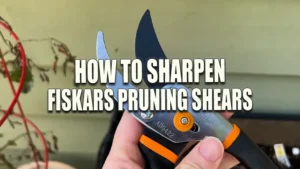 A close-up of a hand holding a pair of open Fiskars Pruning Shears, with a clear view of the sharp blade, against a blurred background of a garden setting.