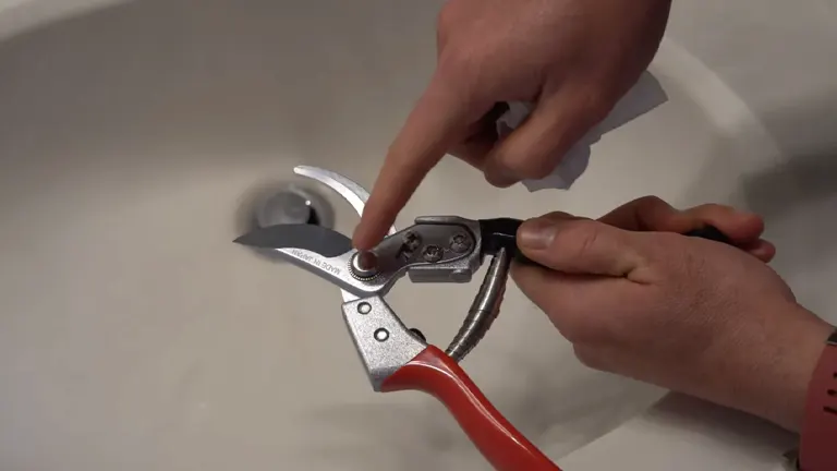 A person's hands are shown adjusting the tension knob of a pair of garden shears with a red handle, over a white basin.