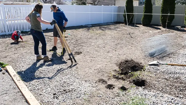 Two people collaborating to measure and mark post locations for a garden fence, with another person digging post holes in the background, in a residential backyard with a roll of wire mesh and gardening tools on the ground.