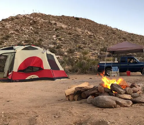 A campsite in a desert setting with a large red and beige tent, a campfire, a blue pickup truck, and a canopy, with a rocky hill in the background.
