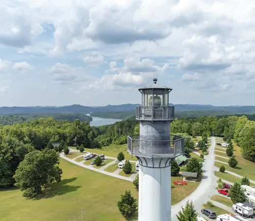 Overlooking a sprawling landscape from a lighthouse viewpoint, with lush trees, a clear view of a distant lake, and an RV park along a winding road under a partly cloudy sky.