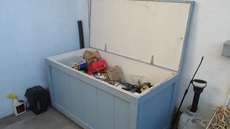 An open, blue outdoor storage box filled with various items, including containers and tools, against a light blue wall, with a pressure washer and other cleaning equipment nearby.