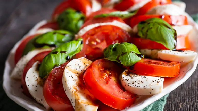 A caprese salad with ripe tomato slices, mozzarella cheese, fresh basil leaves, and a drizzle of balsamic vinegar on a white plate, placed on a wooden table.
