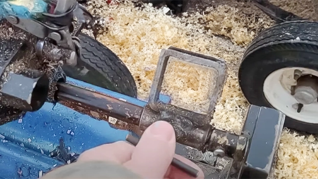 A hand is operating the hydraulic rod of a blue log splitter, with sawdust in the background and a vehicle tire visible