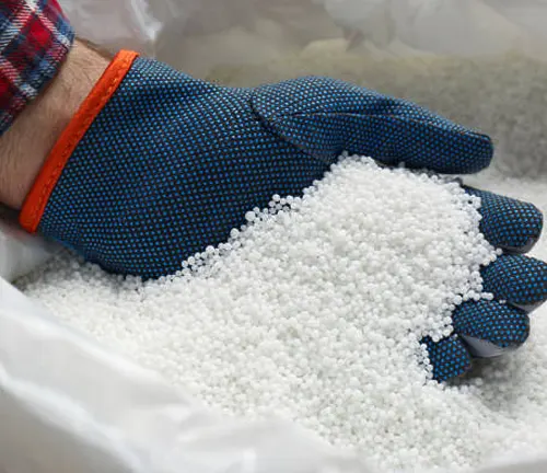 A person wearing a plaid shirt and blue gloves with grip dots holds a handful of white granular fertilizer, suggesting agricultural or gardening activity.