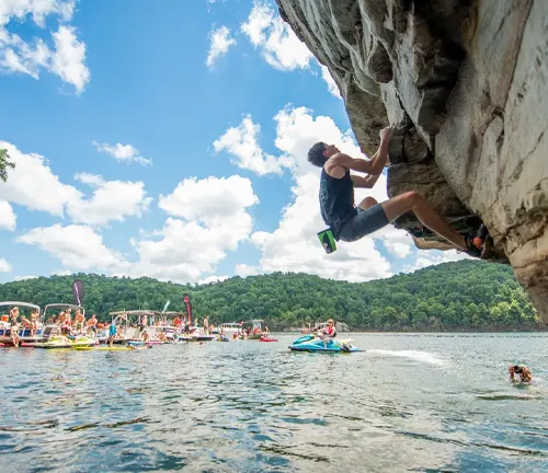 Dynamic view of a rock climber hanging off a cliff above a lively lake scene with boaters, swimmers, and a variety of watercraft under a bright sky with fluffy clouds.