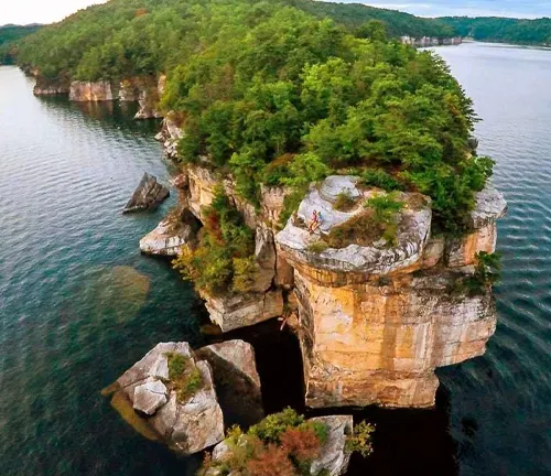An aerial shot capturing the dramatic landscape of a towering cliff adorned with dense greenery, protruding into a vast lake, with a couple standing on the edge, highlighting the grand scale of nature.