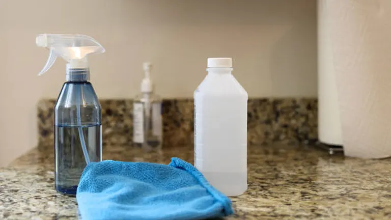 Cleaning supplies on a kitchen countertop including a spray bottle, a white bottle of cleaning solution, hand sanitizer, a blue microfiber cloth, and a roll of paper towels.