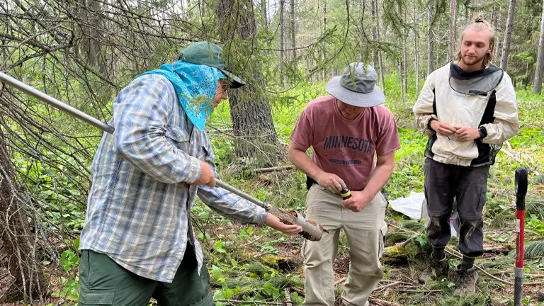 Three people in outdoor attire examining a soil sample in a forested area, with one person holding a soil core sampler and another observing the extracted soil closely.