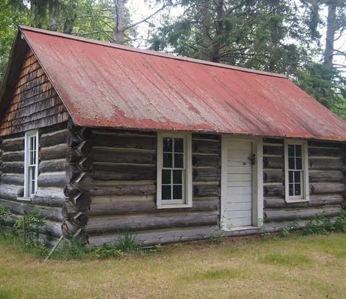 A rustic log cabin with a weathered red roof, nestled among forest trees.