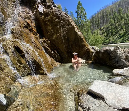 A person relaxes in a clear, natural hot spring at the base of a rocky waterfall surrounded by lush forest.