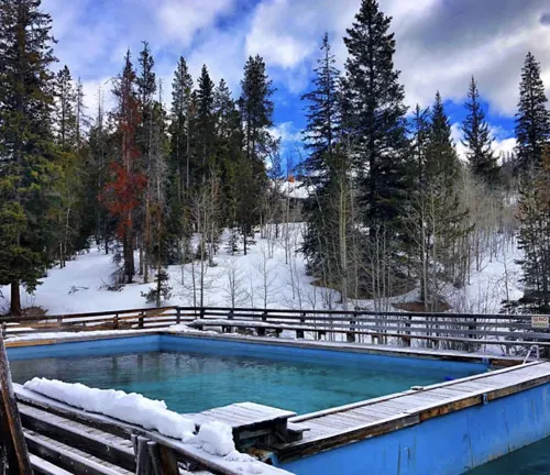 An outdoor hot spring pool surrounded by a wooden fence, with steam rising above the turquoise water, set against a snowy forest landscape and a partly cloudy sky.