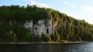 A serene view of a towering limestone cliff draped with dense greenery overlooking calm lake waters under a soft blue sky at dusk.