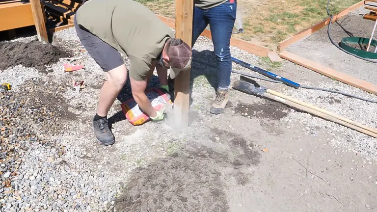  A person in a crouched position pouring concrete mix into a fence post hole, with another person standing by, in a garden area with gravel and soil visible, under clear skies.