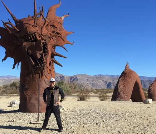 A person posing in front of a large, intricate metal sculpture of a dragon's head with mountains in the distant background.
