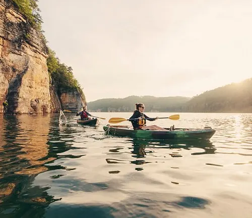 Two kayakers paddle gently on a serene lake with a golden sunset, reflecting on the water near towering cliff walls.