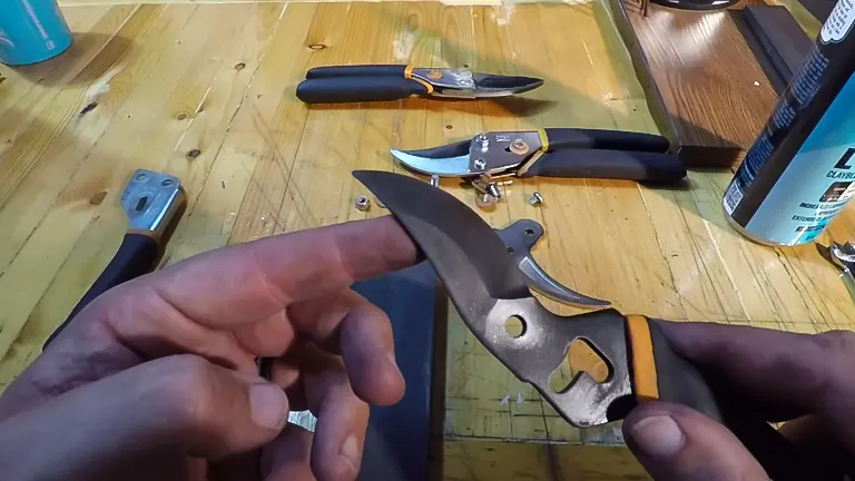 Hands holding a disassembled Fiskars Pruning Shear blade and screw above a wooden table, with the rest of the shear parts and a second assembled pair of shears in the background.