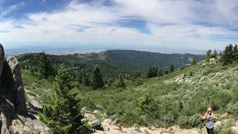Panoramic view from a mountain ridge with large boulders and coniferous trees in the foreground, a woman smiling at the center, and an expansive valley extending towards a hazy horizon under a partly cloudy sky.