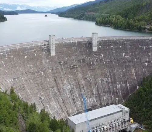 An aerial view of a massive concrete dam, with a hydroelectric power station visible at the base, situated in a forested landscape with a large lake in the background extending into the horizon.