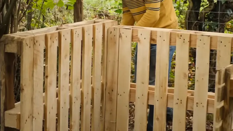 A person in a yellow striped shirt is assembling a wooden compost bin in a garden.