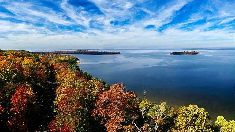 A panoramic autumnal view featuring a forest with a spectrum of fall colors from green to bright red, bordering a tranquil lake under a vast sky with wispy clouds.