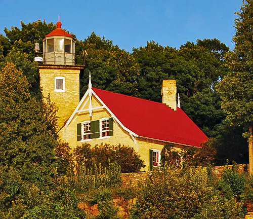 The Eagle Bluff Lighthouse with its classic stone structure and red roof, nestled among green trees under a clear sky, stands as a historical beacon on the shores of Peninsula State Park.