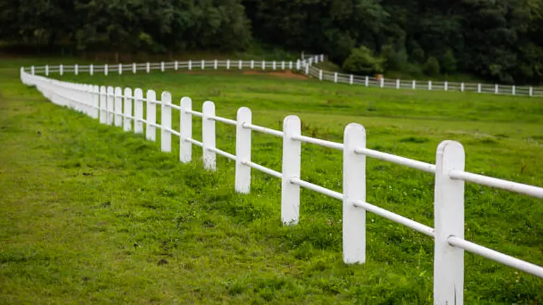 A neat white wooden fence with two horizontal slats on a lush green lawn, next to a puddle of water and some scattered debris on the ground, suggesting recent rainfall.
