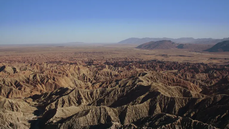 Aerial view of a vast, rugged canyon with intricate erosion patterns, extending towards a flat horizon with distant mountains under a hazy sky.