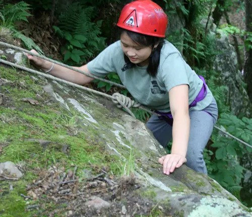 A person in a helmet climbing a mossy rock with ropes for support.

