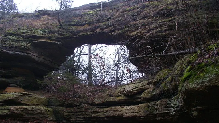 Dimly lit view of a smaller natural rock arch in a forested area, with bare trees and a trail leading underneath the arch.